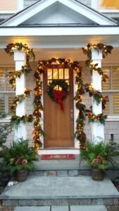 wreath with a bow on exterior door, framed by garland and string lights wrapped around railing and pillars, illuminated in the day